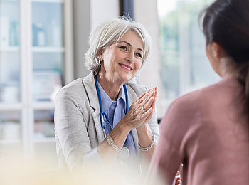 Eine ältere Hausärztin mit kurzen grauen Haaren führt ein Arztgespräch mit einer Patientin. Sie lächelt dabei freundlich.