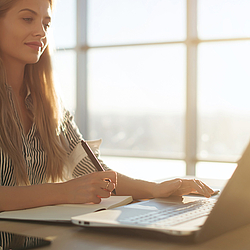 Eine Frau mit langen blonden Haaren sitzt an einem Tisch. Vor ihr steht ein Laptop und sie schreibt in ein Notizbuch.