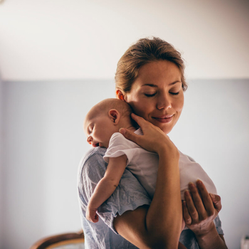 Ein junge Mutter hat ihr Neugeborenes auf ihre Schulter gelegt. Eine Hand hat sie am Rücken des Babys, die anderen unter seinem Po. Sie hat die Augen geschlossen und lächelt.