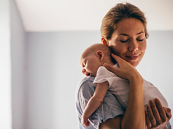 Ein junge Mutter hat ihr Neugeborenes auf ihre Schulter gelegt. Sie hat die Augen geschlossen und lächelt.