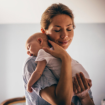 Ein junge Mutter hat ihr Neugeborenes auf ihre Schulter gelegt. Sie hat die Augen geschlossen und lächelt.