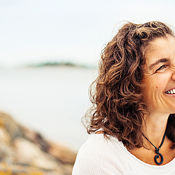 Eine Frau mit lockigen Haaren steht in der Natur. Sie trägt ein weißes Shirt und lacht ausgelassen.