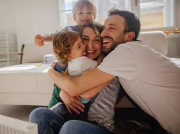 Eine junge vierköpfige Familie sitzt auf dem Boden vor ihrem Sofa im Wohnzimmer. Alle lachen und umarmen sich innig.
