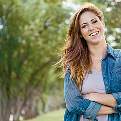 Eine junge Frau mit langen braunen Haaren steht in der Natur. Sie trägt ein Jeanshemd und lacht fröhlich in die Kamera.
