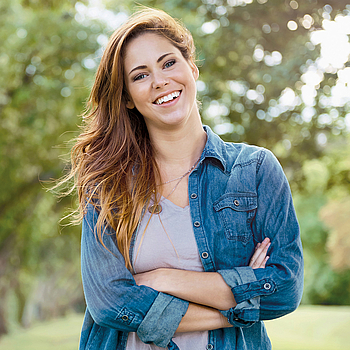 Eine junge Frau mit langen braunen Haaren steht in der Natur. Sie trägt ein Jeanshemd und lacht fröhlich in die Kamera.