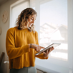 Eine Frau steht vor dem Fenster und hat ein Tablet in der Hand. Sie lächelt.