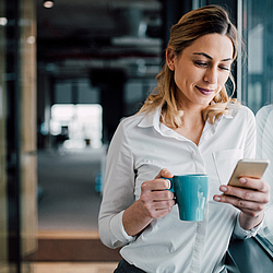 Eine Frau mit langen blonden Haaren lehnt an einem Fenster. Sie trägt eine weiße Bluse, schaut auf ein Smartphone und lächelt.