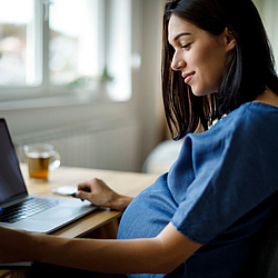 Eine schwangere Frau mit dunklen Haaren sitzt an einem Schreibtisch. Vor ihr steht ein Laptop, daneben liegen Dokumente, die sie ausfüllt.