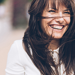 Eine Frau mit langen braunen Haaren lacht ausgelassen in die Kamera. Der Wind weht ihr Haare ins Gesicht.