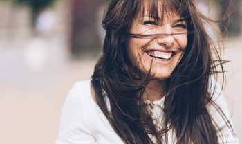 Eine junge Frau mit langen braunen Haaren lacht ausgelassen in die Kamera. Der Wind weht ihr Haare ins Gesicht.