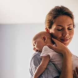 Eine Mutter hält ihr Neugeborenes auf dem Arm und lächelt dabei. Das Baby liegt schlafend auf ihrer Schulter.