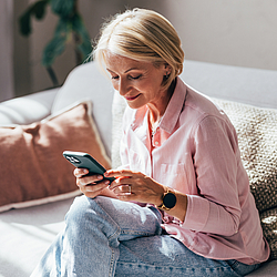 Eine Frau in einer rosafarbenen Bluse sitzt auf einem Sofa. Sie schaut auf ihr Smartphone.