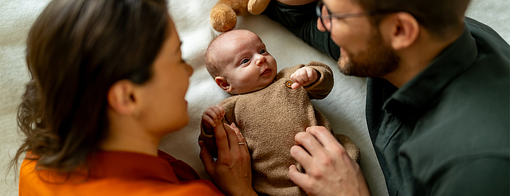 Mutter und Vater liegen mit ihrem Baby auf einer Decke. Sie lächeln das Baby an.