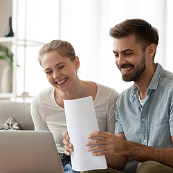 Ein junges Paar sitzt lächelnd auf dem Sofa. Sie schauen auf einen Laptop und lächeln. Der Mann hält Papiere in der Hand.