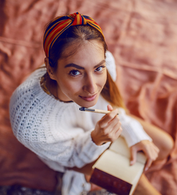 Eine junge Frau mit langen Haaren und einem bunten Tuch im Haar, sitzt auf einer roten Decke. Sie trägt einen weißen Pullover, auf ihrem Schoß liegt ein Notizbuch. In ihrer Hand hält sie einen Stift. Sie schaut lächelnd nach oben in die Kamera.