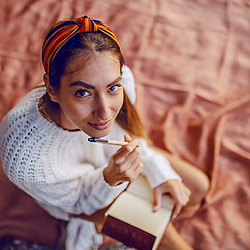 Eine Frau mit langen Haaren und einem bunten Tuch im Haar, sitzt auf einer roten Decke. Auf ihrem Schoß liegt ein Notizbuch.