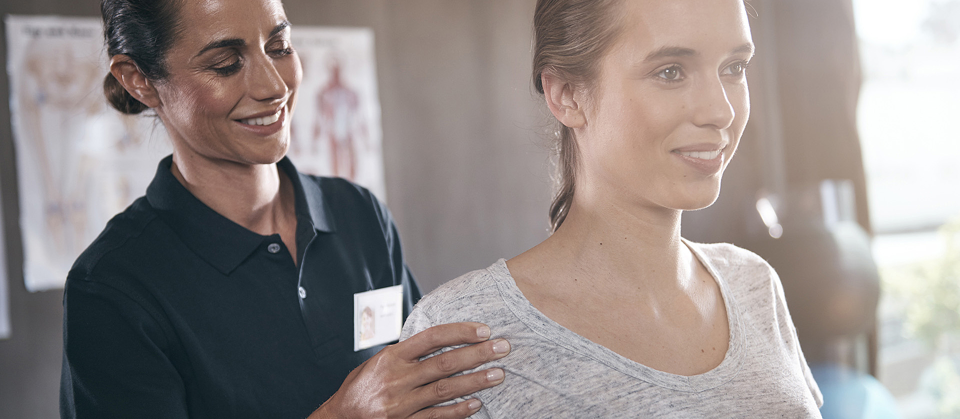 Eine junge lächelnde Frau bekommt Physiotherapie. Die lächelnde Therapeutin steht hinter ihr und fasst ihr an die Schulter.