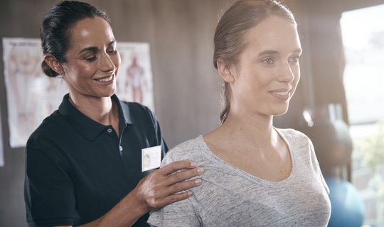 Eine junge lächelnde Frau bekommt Physiotherapie. Die lächelnde Therapeutin steht hinter ihr und fasst ihr an die Schulter.