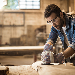 Ein junger Tischler arbeitet in seiner Werkstatt mit einem Schleifgerät. Er trägt eine Schutzbrile und Handschuhe.