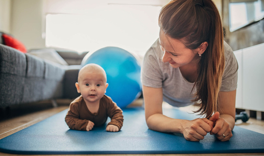 Eine junge Mitter mit langen dunklen Haaren liegt in Sportkliedung auf einem Gymnastikmatte. Sie schaut lachend auf ihr Baby, das neben ihr auf der Mattte liegt. Das Baby schaut fröhlich in die Kamera.