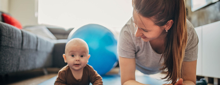 Eine junge Mitter mit langen dunklen Haaren liegt in Sportkliedung auf einem Gymnastikmatte. Sie schaut lachend auf ihr Baby, das neben ihr auf der Mattte liegt. Das Baby schaut fröhlich in die Kamera.