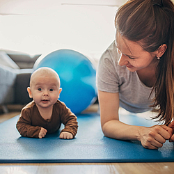 Eine junge Mitter liegt in Sportkleidung auf einem Gymnastikmatte. Sie schaut lachend auf ihr Baby, das neben ihr auf der Mattte liegt. 