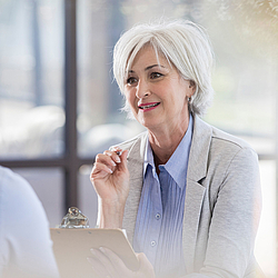 Eine Frau mit kurzen grauen Haaren hält einen Stift und ein Klemmbrett in den Händen. Sie ist im Gespräch mit einem Mann.