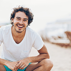 Ein Mann in einem weißen Shirt lächelt. Im Hintergrund ist Strand zu sehen.