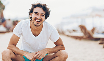 Ein Mann in einem weißen Shirt lächelt. Im Hintergrund ist Strand zu sehen.
