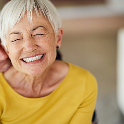 Eine ältere Frau mir kurzen weißen Haaren lacht. Ein älterer Mann umarmt sie lächelnd von hinten und legt den Kopf auf ihre Schulter.