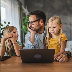 Ein Vater mit kurzen Haaren sitzt mit seinen Töchtern an einem Tisch. Ein Mädchen schaut ihren Vater an und erzählt ihm etwas