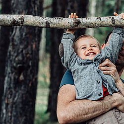Ein Vater hebt seinen Sohn im Wald hoch, damit der Sohn an einem Ast hängen kann. Sie lachen beide dabei.