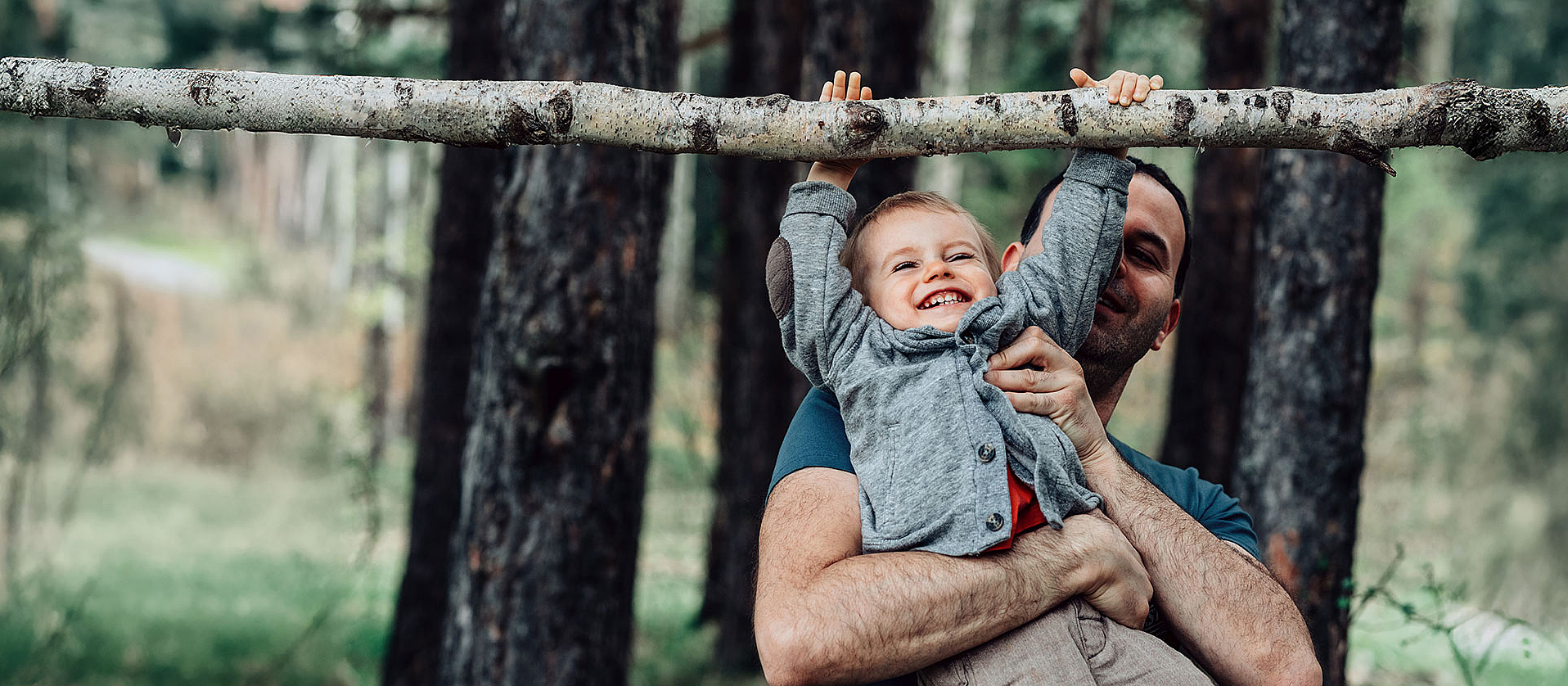 Ein Vater hebt seinen Sohn im Wald hoch, damit der Sohn an einem Ast hängen kann. Sie lachen beide dabei.