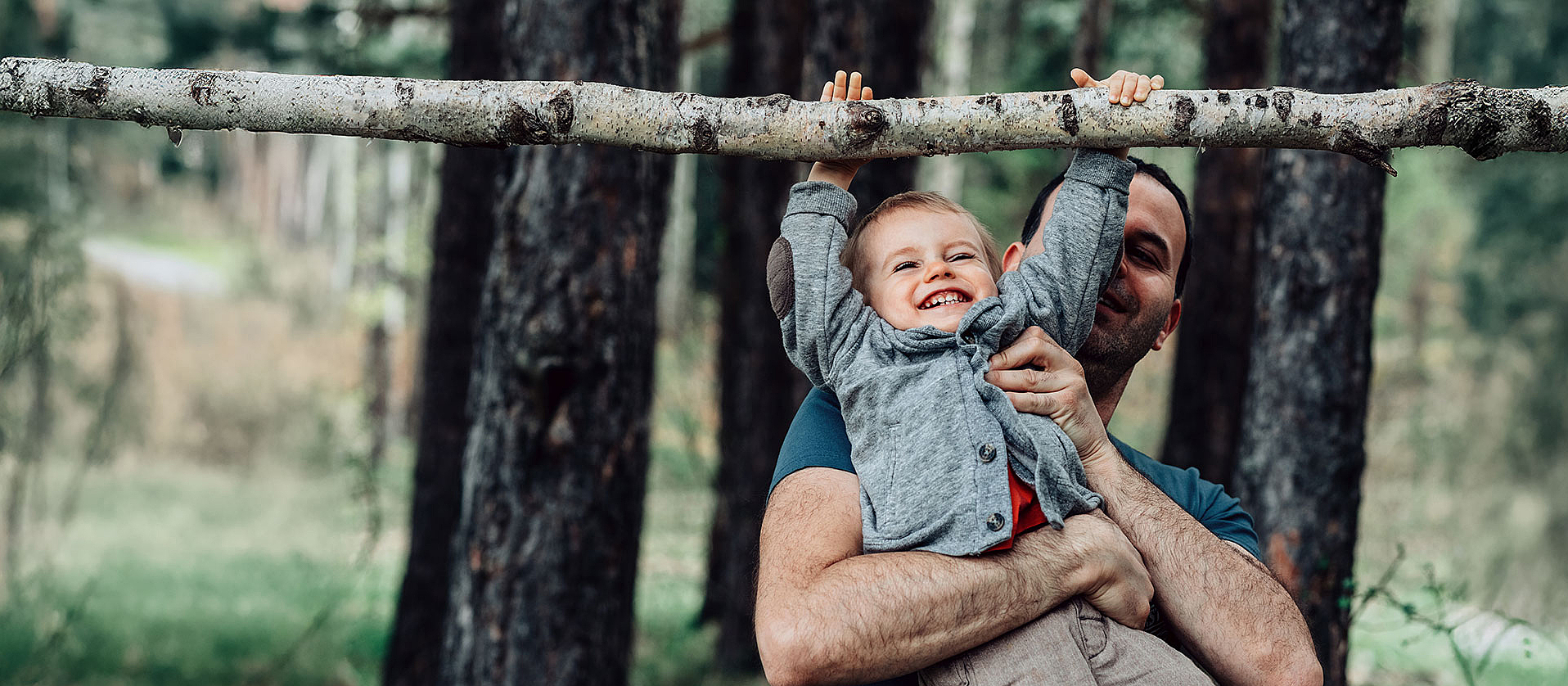 Ein Vater hebt seinen Sohn im Wald hoch, damit der Sohn an einem Ast hängen kann. Sie lachen beide dabei.