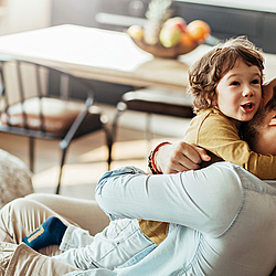 Ein Vater mit dunklen Haaren und Bart sitz auf einem Sofa. Er hat seinen kleinen Sohn auf dem Arm und drückt ihn fest an sich.