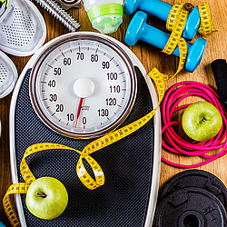 Um eine analoge Waage herum liegen Hanteln, ein Maßband, Äpfel, ein Springseil, eine Yogamatte, eine Schale mit Müsli und ein Paar Turnschuhe.