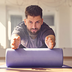 Ein Mann mit Bart sitzt auf einer Trainingsmatte und macht Übungen mit einer Faszienrolle.