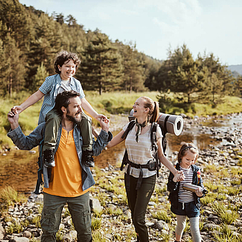 Eine vierköpfige Familie wandert an einem Fluss entlang. Im Hintergrund sieht man einen Wald. Der Sohn sitzt auf den Schulter vom Vater, alle halten sich an den Händen und lachen ausgelassen.