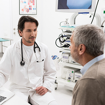 Ein Arzt sitzt am Laptop und spricht mit seinem älteren Patienten über die Darmkrebsvorsorge.