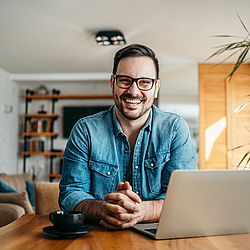 Ein Mann mit Bart und Brille sitzt an einem Tisch und schaut lachend in die Kamera. Vor ihm stehen ein Laptop und eine Kaffeetasse.