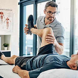 Ein Mann liegt auf einer Liege beim Physiotherapeuten. Der Therapeut hält sein Bein angewinkelt hoch und drückt aufs Knie.