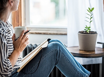 Eine Frau schaut aus dem Fenster. Auf Ihrem Schoß liegt ein aufgeschlagenes Notizbuch. In der Hand hält sie eine Tasse.
