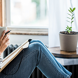 Eine Frau schaut aus dem Fenster. Auf Ihrem Schoß liegt ein aufgeschlagenes Notizbuch. In der Hand hält sie eine Tasse.