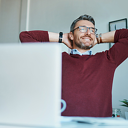Ein Mann mit leicht grauen Haare, Bart und Brille sitzt an einem Tisch. Vor ihm stehen ein Laptop und eine Tasse.
