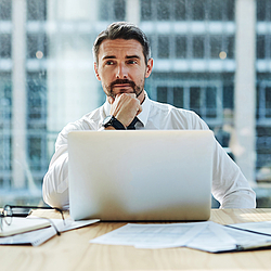 Ein Mann mit dunklen Haaren sitzt an einem Tisch. Vor ihm steht ein Laptop, daneben liegen Dokumente. Der Mann schaut nachdenklich. 