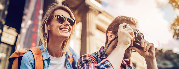 Auslandskrankenschein Junge Frau mit Sonnenbrille und Mann mit Kamera schauen in die Ferne