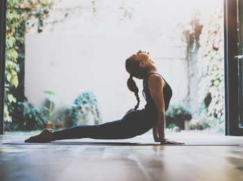 Eine junge Frau mit langem geflochtenen Zopf macht eine Yoga-Pose am geöffneten Fenster.
