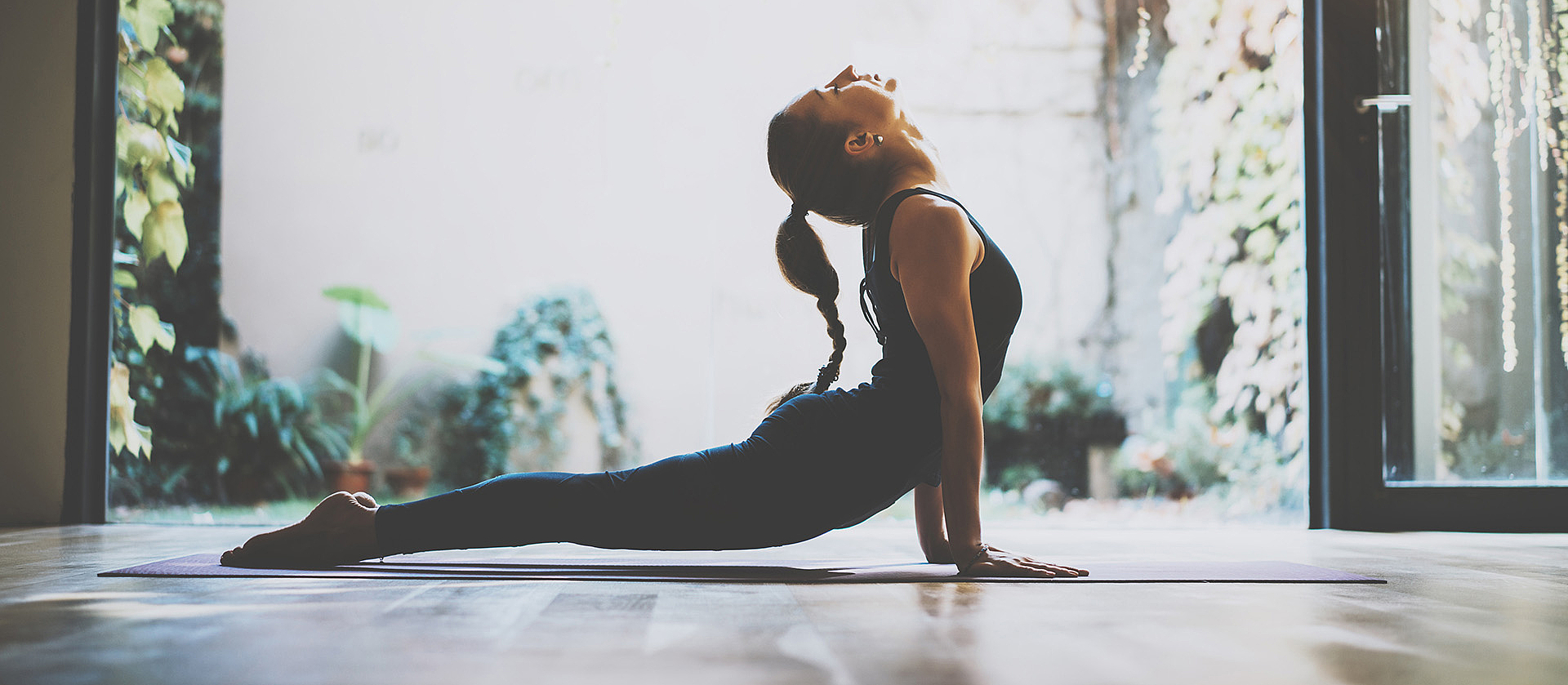 Eine junge Frau mit langem geflochtenen Zopf macht eine Yoga-Pose am geöffneten Fenster.