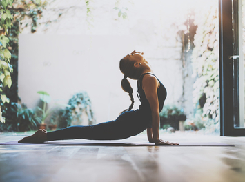 Eine junge Frau mit langem geflochtenen Zopf macht eine Yoga-Pose am geöffneten Fenster.