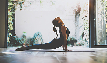 Eine junge Frau mit langem geflochtenen Zopf macht eine Yoga-Pose am geöffneten Fenster.